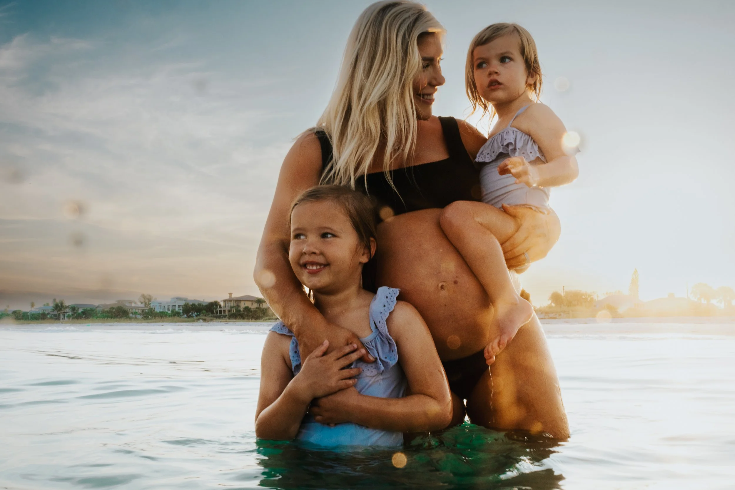 A woman and three young girls enjoying a moment in the ocean during sunset, with houses visible in the background.
