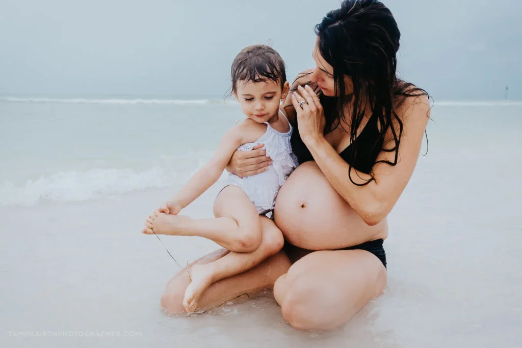 A pregnant woman and a young girl sitting on the beach with gentle waves in the background.