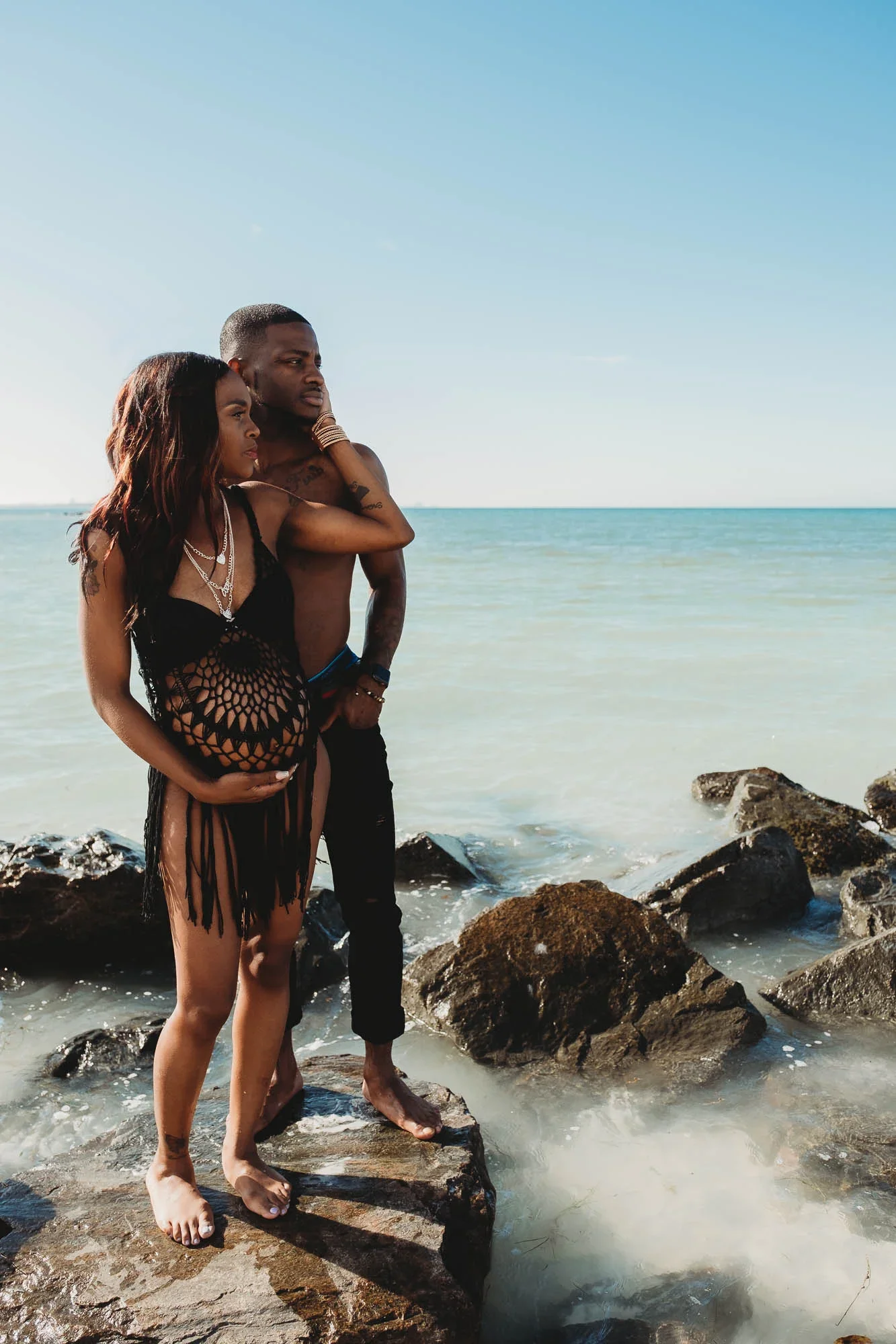 A couple standing on rocks at the beach near the water, with the ocean and clear sky in the background.