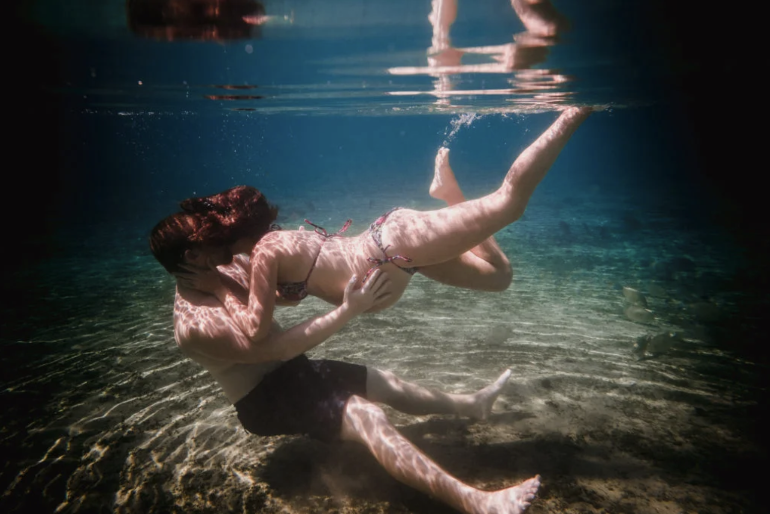 A woman underwater in a bikini, embracing and kissing her partner in a shallow, clear body of water.