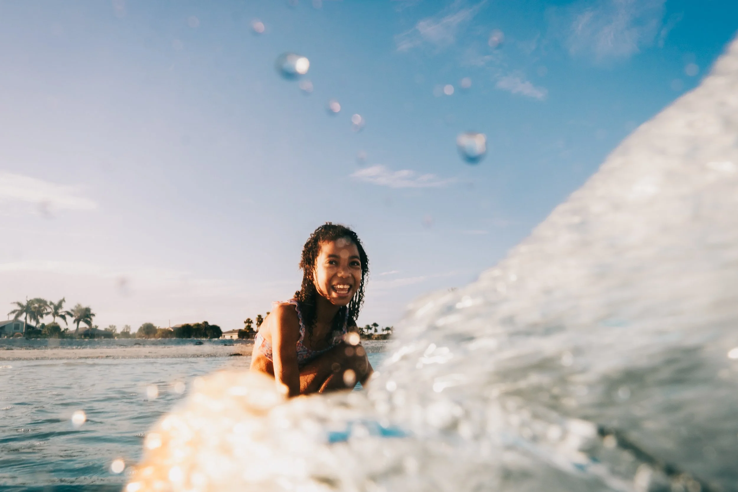 A person smiling at the camera while sitting in the ocean near the shoreline during daytime, with water splashing and a blue sky with some clouds in the background.