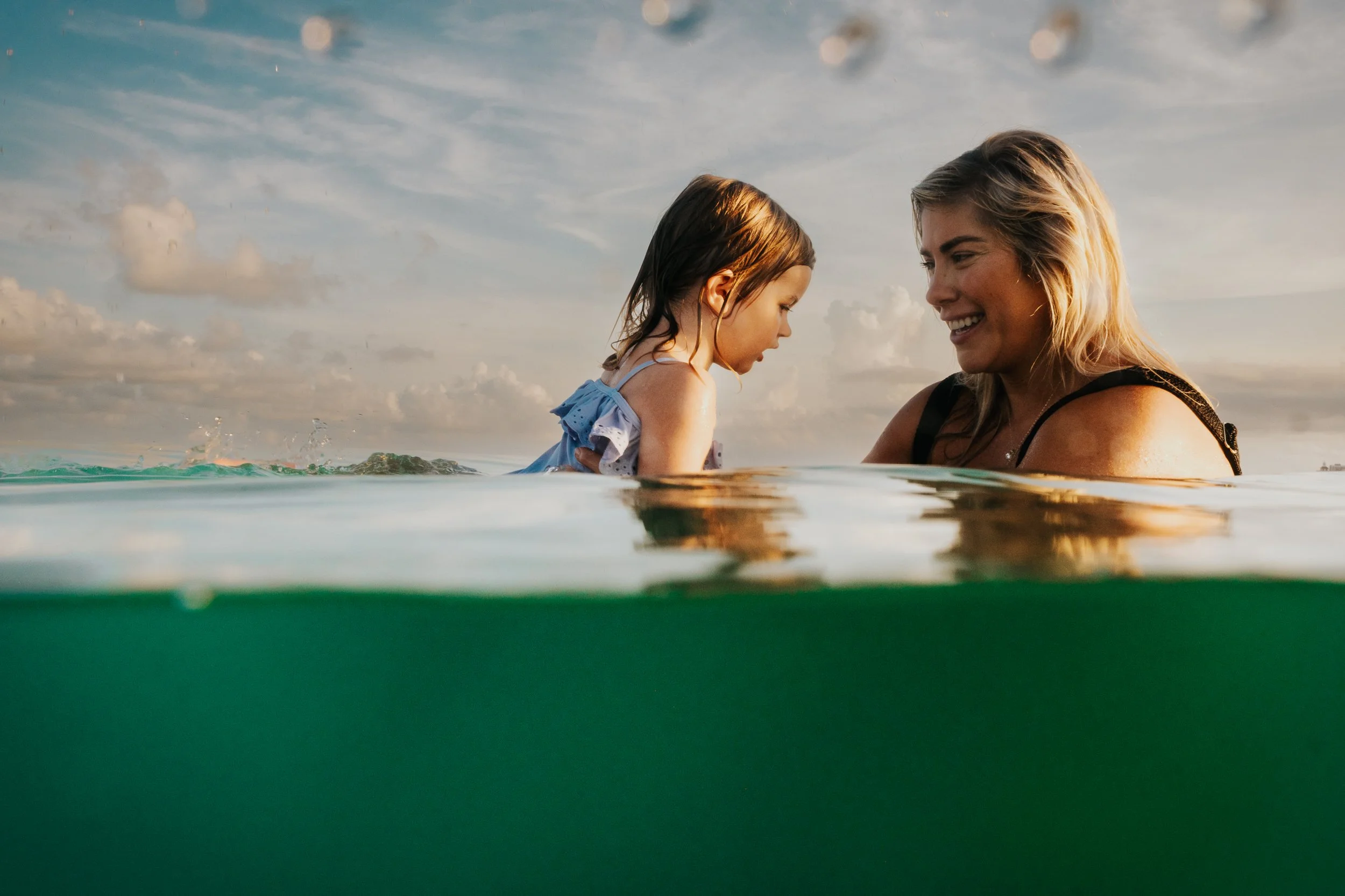 A woman and a young girl are smiling and enjoying a swim in the ocean at sunset, with the photo taken partially underwater.