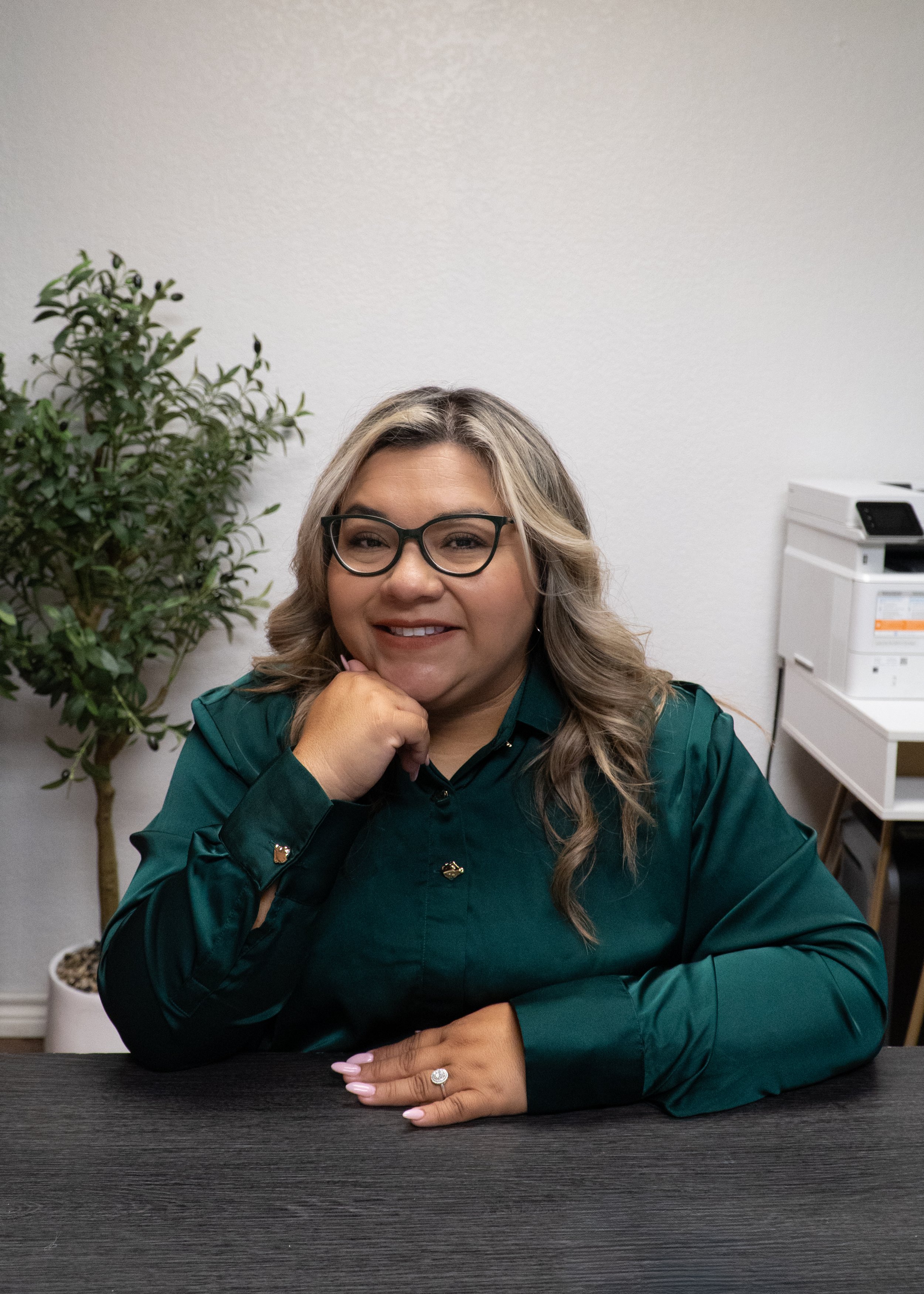 A woman with long wavy blonde hair, glasses, and a ring on her left hand, smiling and resting her chin on her hand, sitting at a dark wood table in an office or conference room, with a white wall, green plant, and a white filing cabinet in the background.