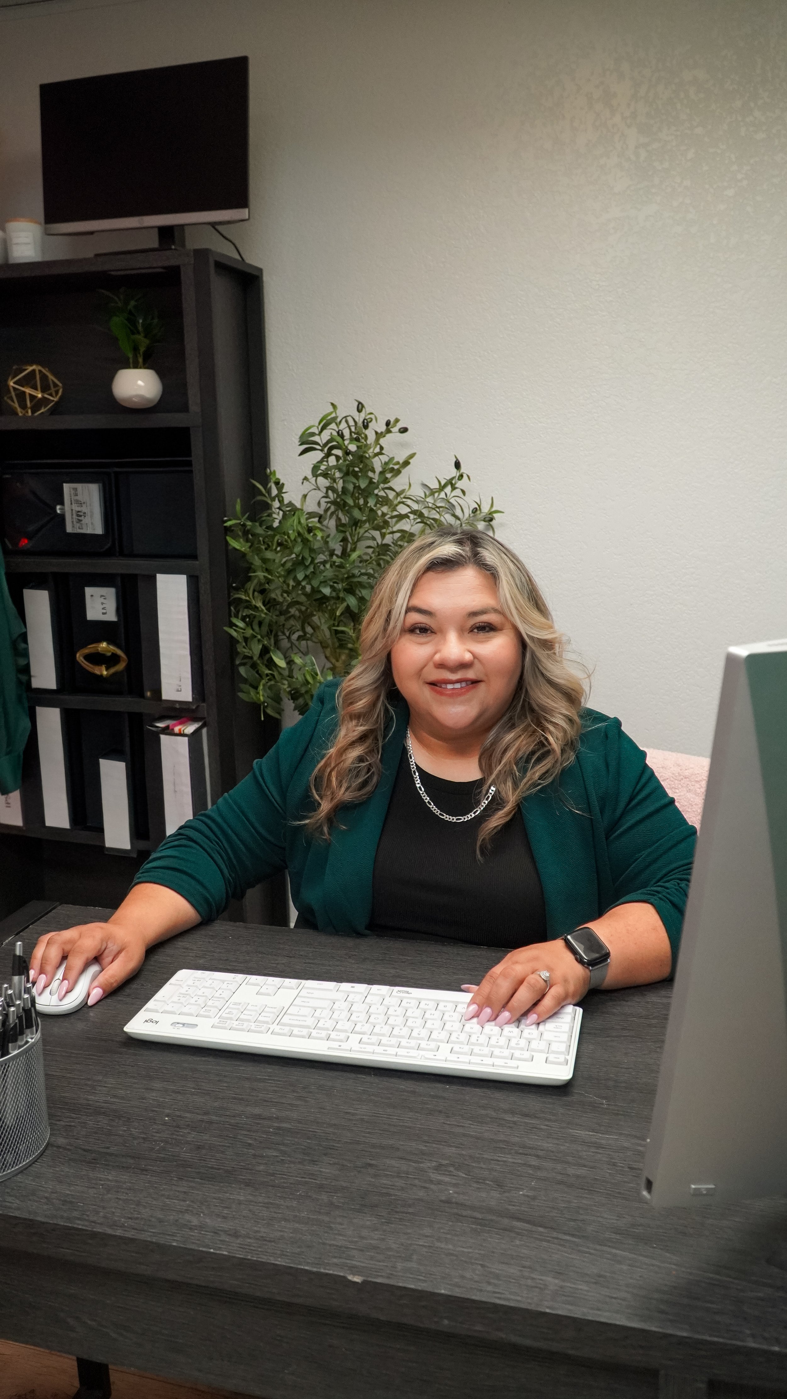 A woman with wavy blonde hair, wearing a black top, green blazer, and silver necklace, sitting at a desk with a computer keyboard, mouse, pens in a holder, and a monitor, smiling at the camera in an office setting with a white wall, bookshelf, and green plant behind her.
