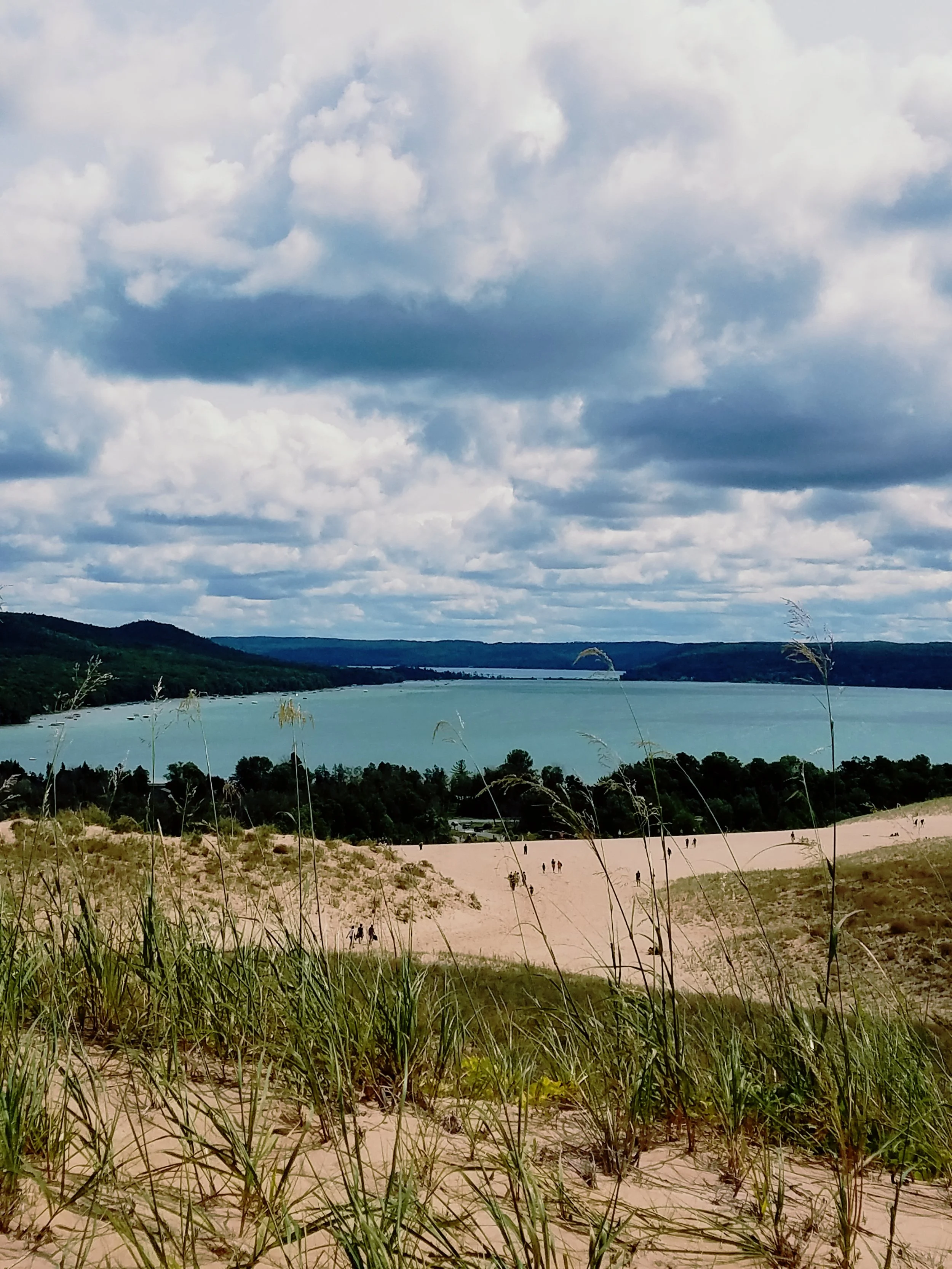 View of a large body of water, likely a lake or river, with hills or mountains in the background. The sky is partly cloudy with large clouds, and there is a sandy area with some people walking and grass in the foreground.