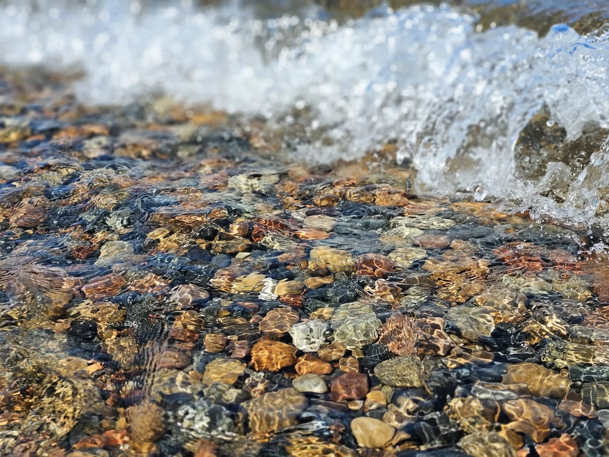 Close-up of small multicolored pebbles in clear, shallow water, with a small splash of water at the top right.