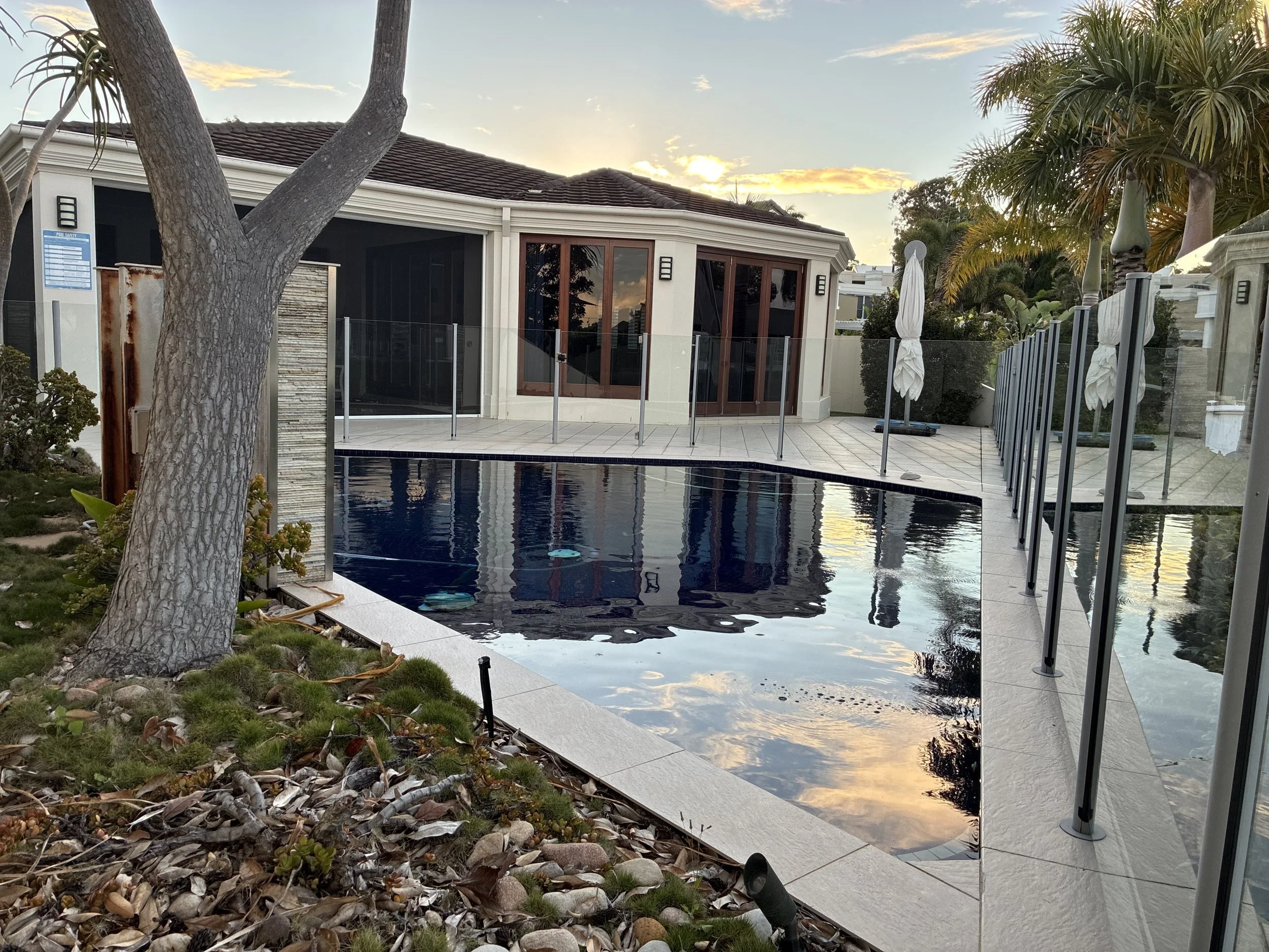 A backyard with a swimming pool, a patio area with glass fencing, and a house with large windows and a dark roof. There are palm trees and a sunset in the background.