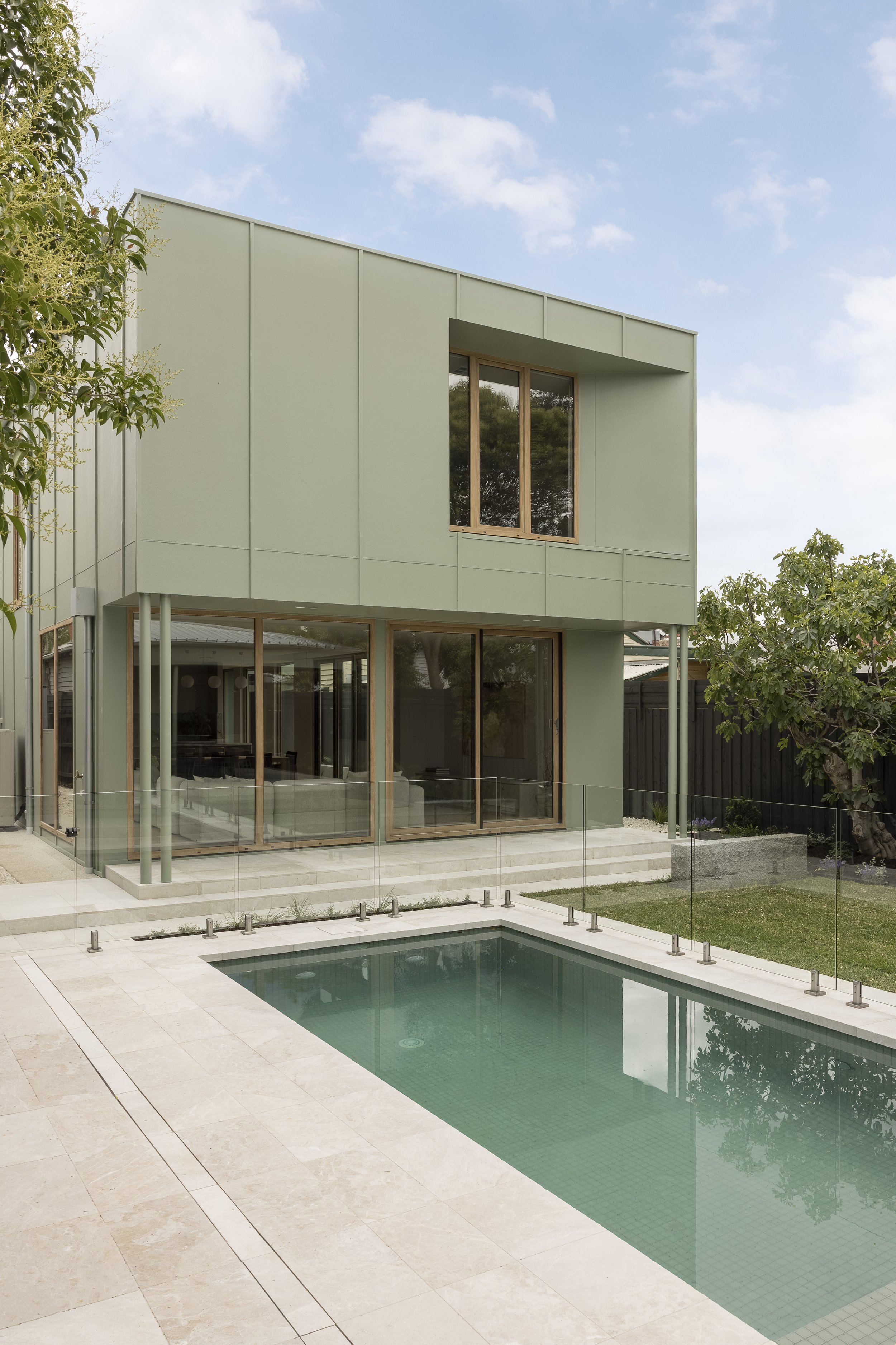 Modern house with large windows, outdoor pool, and a glass fence, surrounded by trees, under a partly cloudy sky.