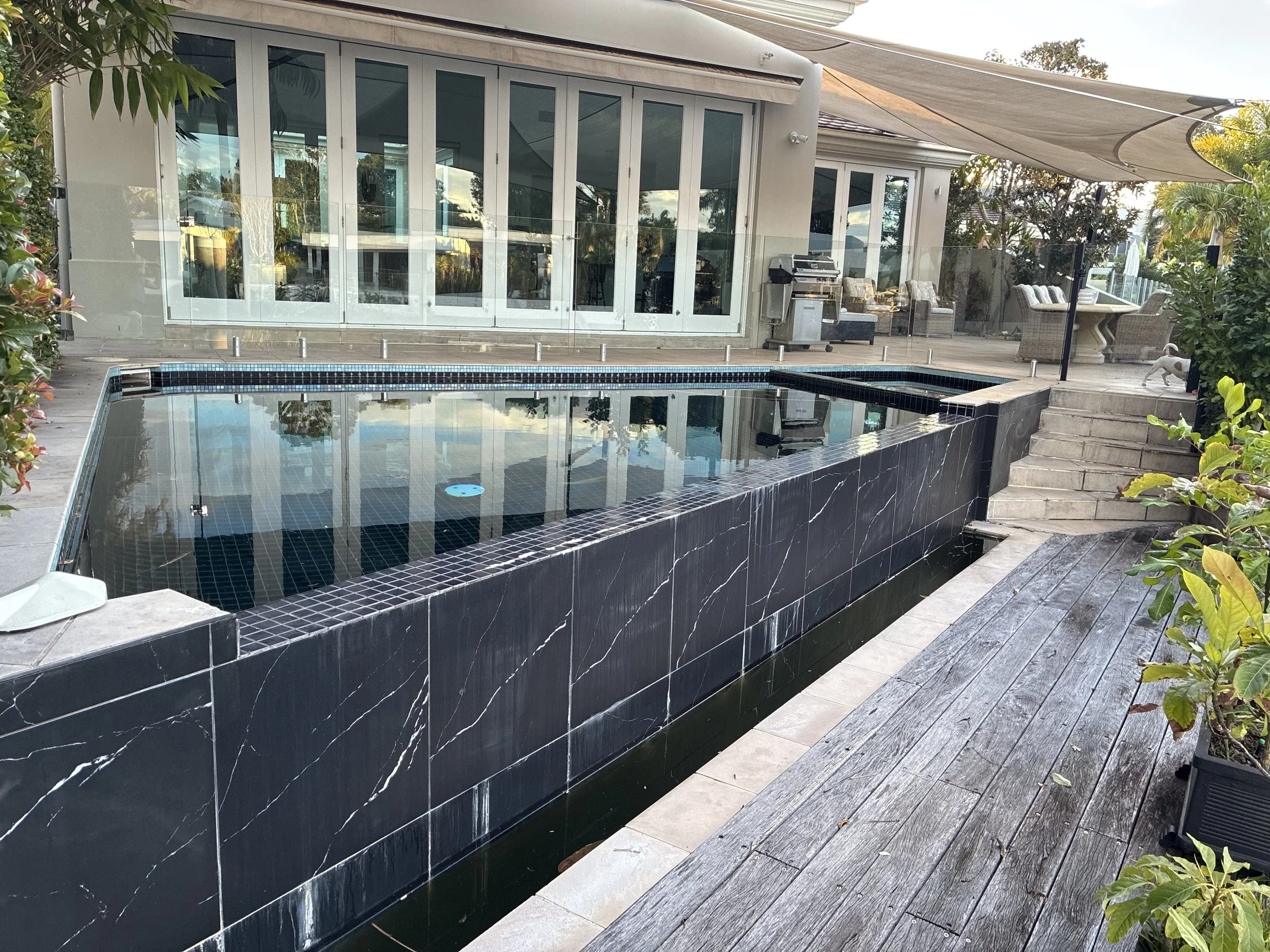 A backyard with a swimming pool, outdoor furniture, and a detached house with large glass windows and sliding doors. There is a shaded area with a canopy and potted plants.