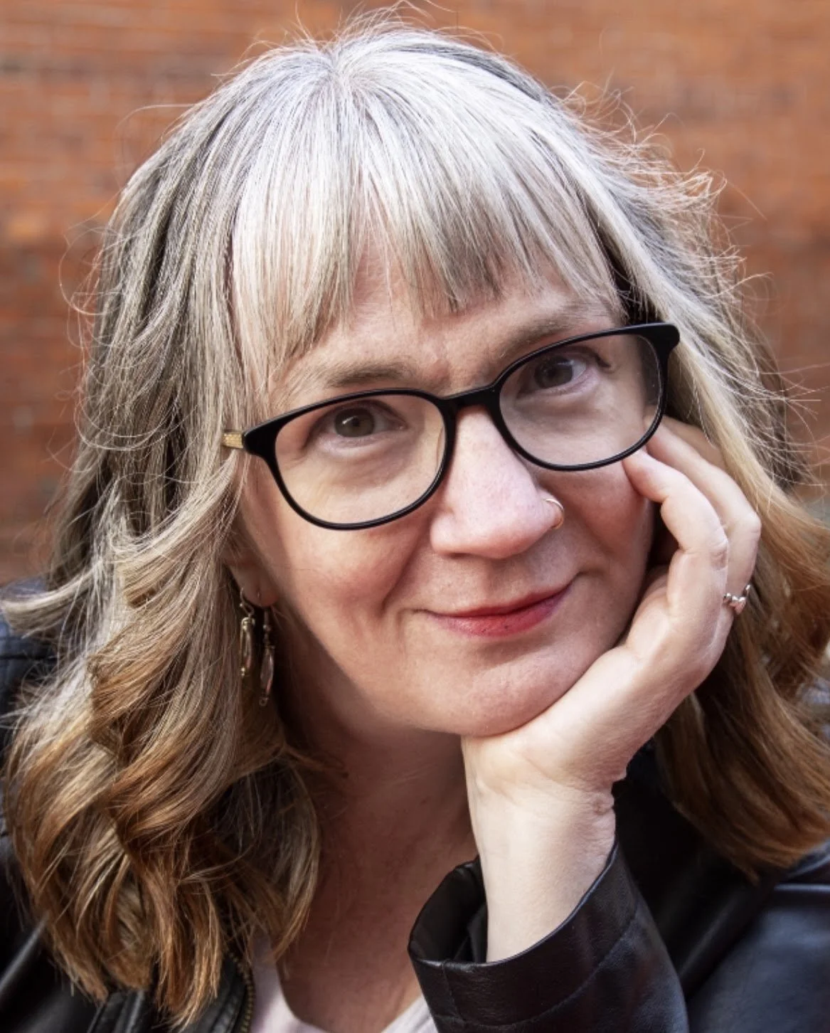 Close-up of a woman with long, wavy blonde hair and glasses, resting her face on her hand, outdoors with a brick wall background.