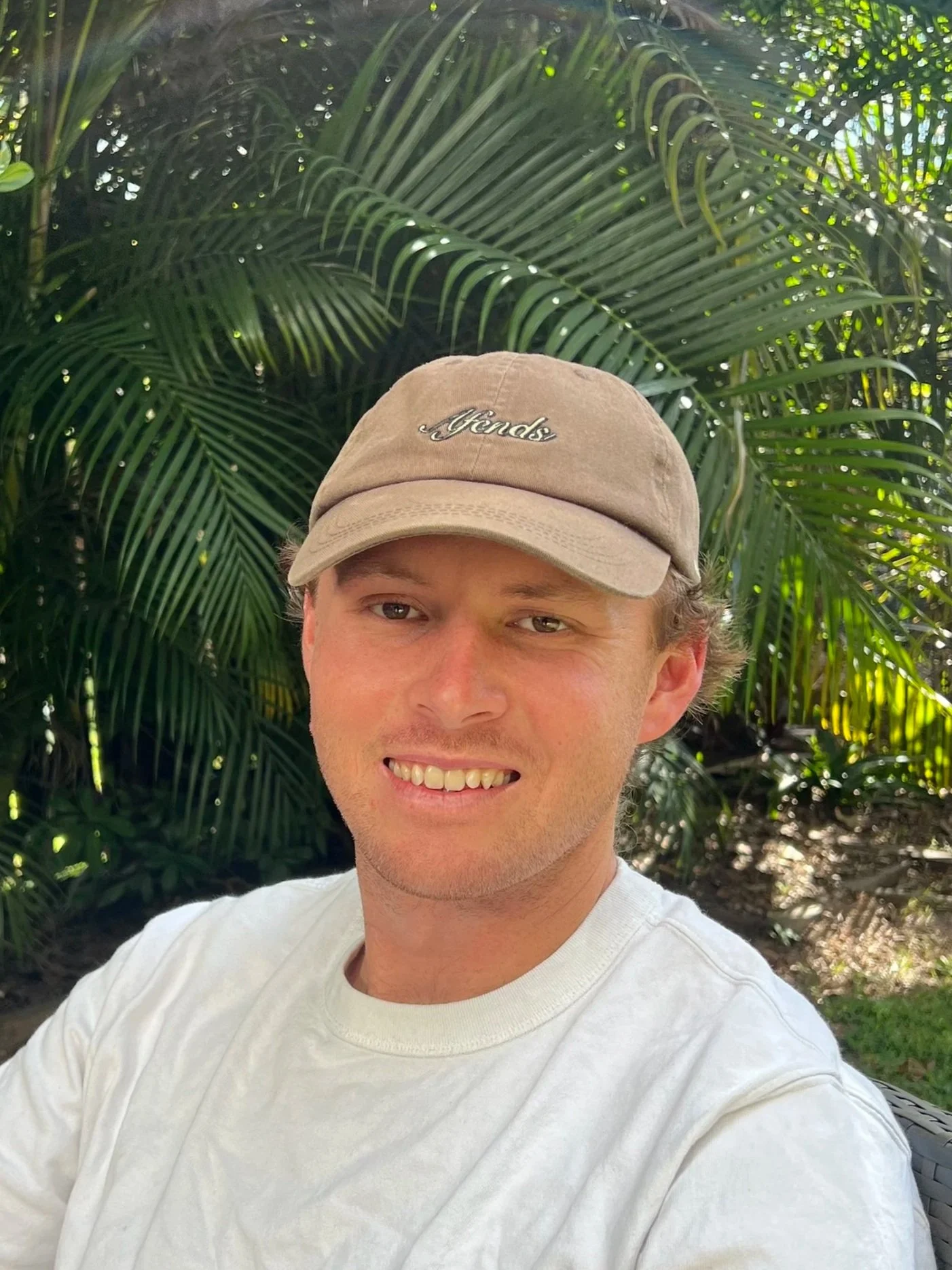 A young man smiling, wearing a beige cap with the word 'Fendi' embroidered on it, outdoors with lush green tropical plants in the background.