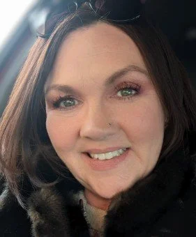 Close-up of a woman with shoulder-length brown hair, smiling, wearing makeup, and a dark fur-like coat.