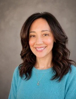 A woman with dark hair dressed in a blue sweater, smiling with a neutral gray background.
