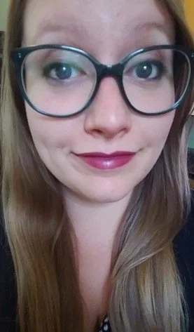Close-up of a young woman with glasses, long light brown hair, and pink lipstick, smiling slightly.