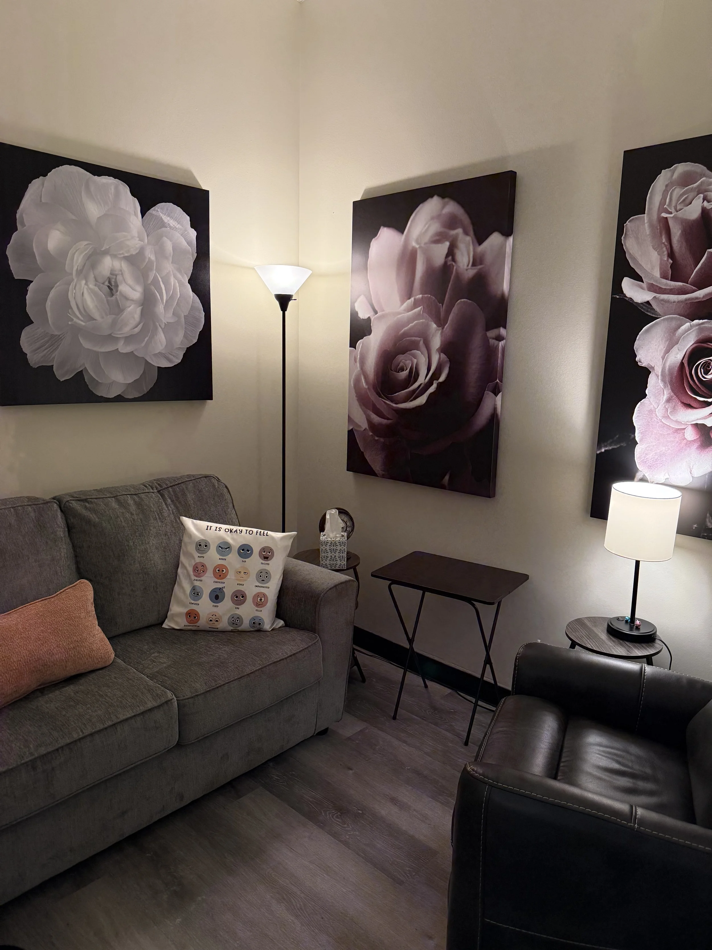 Living room with a gray sofa, black leather chair, framed flowers on the wall, a lamp, a side table, and decorative pillows.