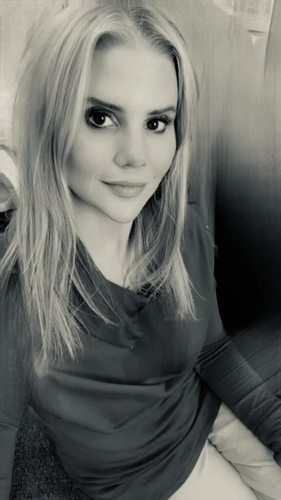 A black and white photo of a young woman with long, straight blonde hair, smiling slightly, sitting indoors against a wooden wall.