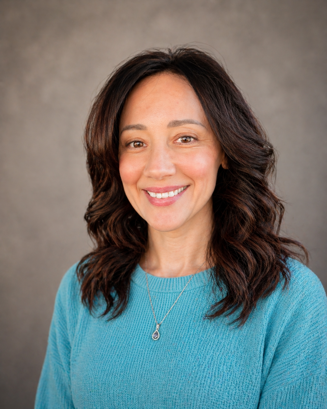 Headshot of a smiling woman with wavy dark hair, wearing a turquoise sweater and a silver necklace, against a neutral background.