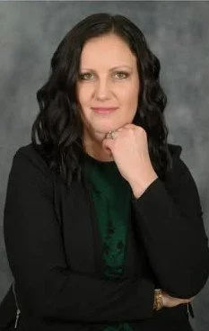 Professional portrait of a woman with dark wavy hair, wearing a black blazer and green top, posed with her hand near her chin against a gray backdrop.
