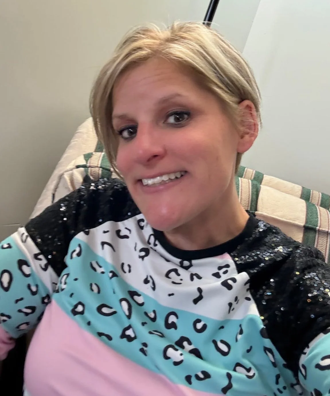 A woman with short blonde hair taking a selfie, sitting on a beige and green striped couch, wearing a colorful shirt with black sequin details.