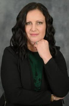 A woman with shoulder-length wavy black hair, wearing a dark jacket over a green top, posing with her hand resting on her chin against a gray background.