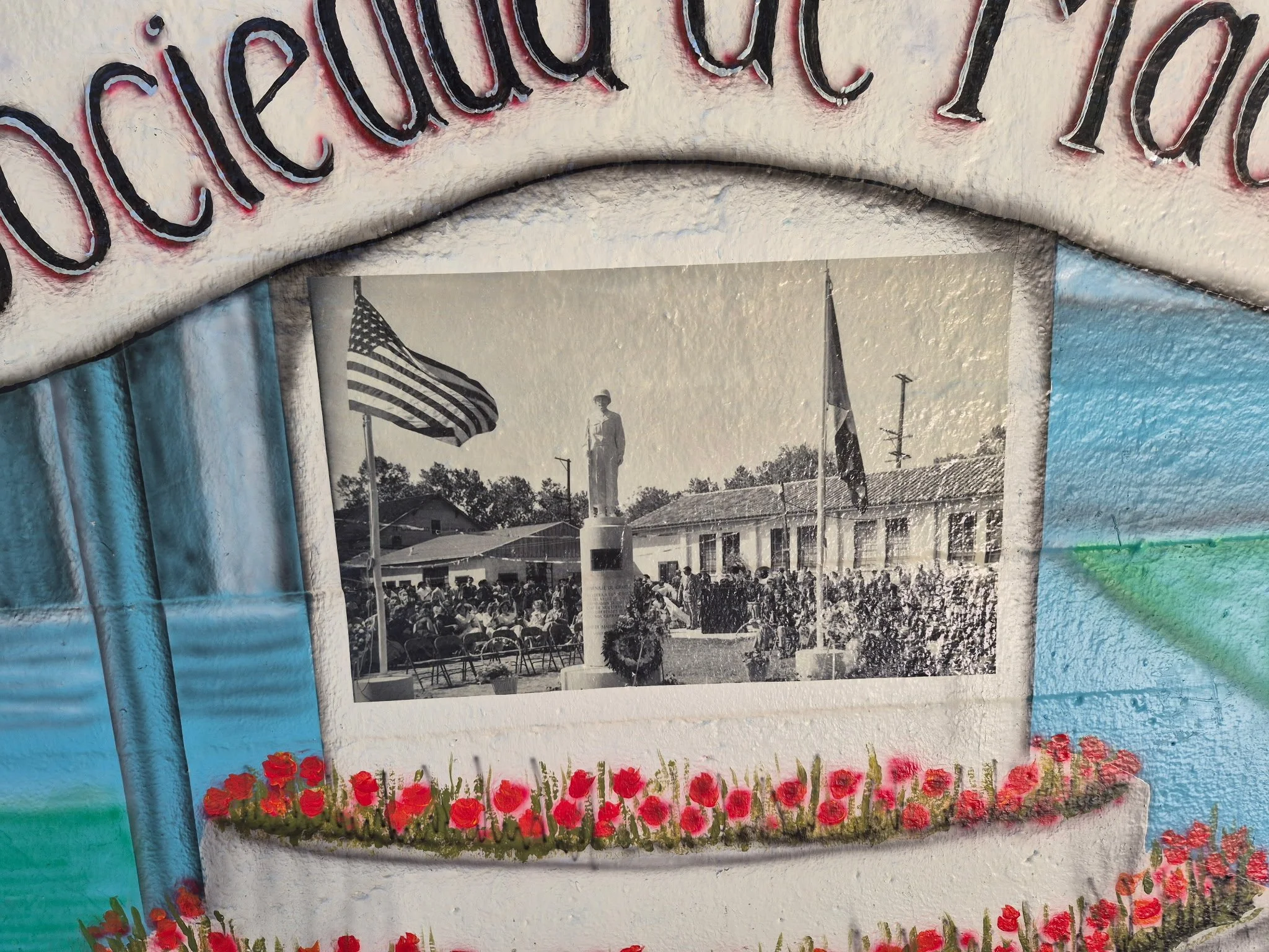 A black and white photograph of a crowd gathered in front of a monument with flags, surrounded by a mural with red flowers at the bottom.