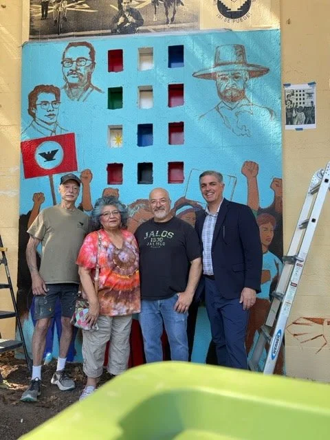 Four people, two men and two women, stand in front of a large mural depicting portraits of historical figures and protest artwork, with a ladder to the right.