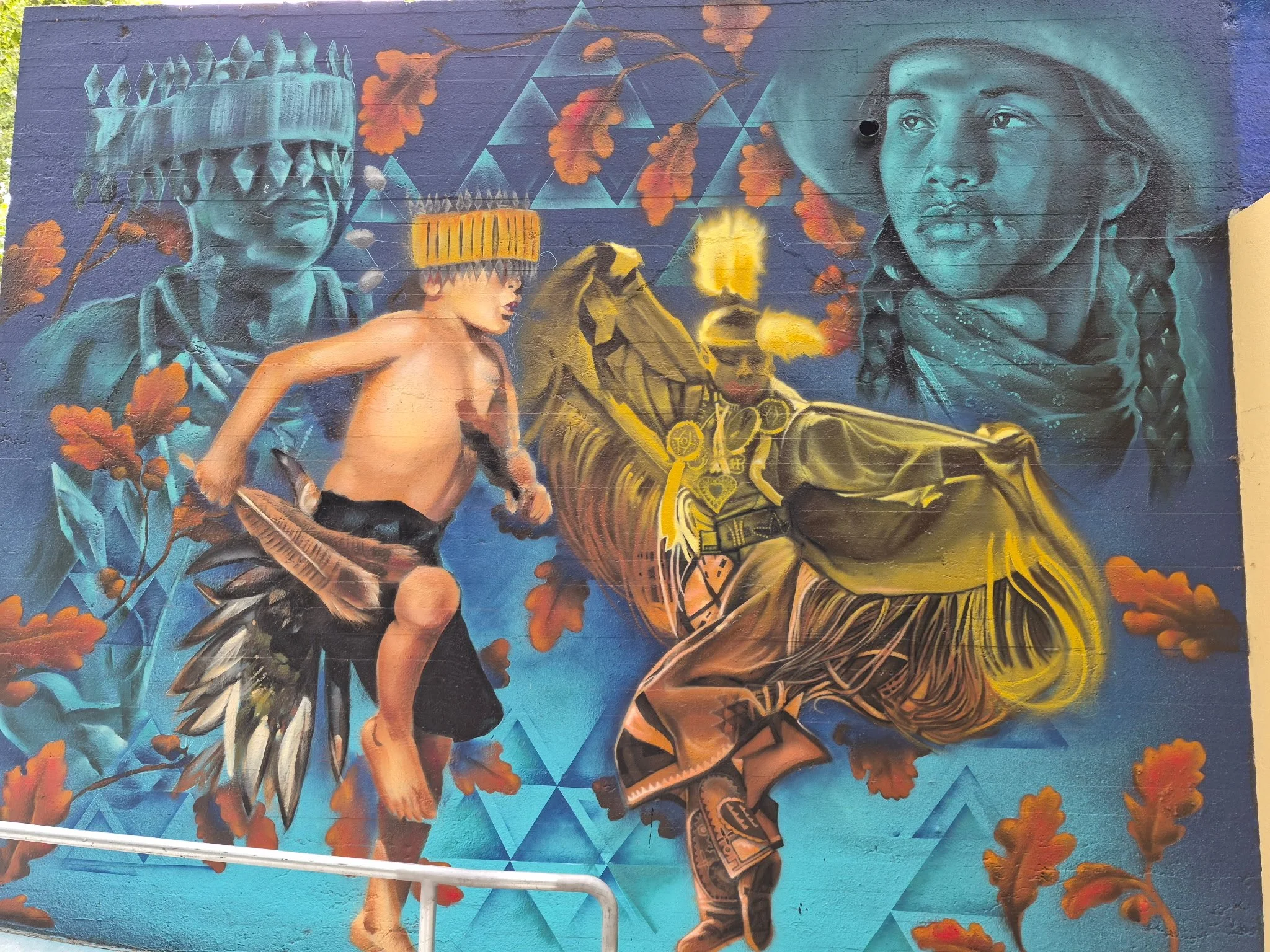 Colorful mural depicting two women, one holding a bow and arrow, and the other wearing a traditional headdress, with vibrant autumn leaves and geometric patterns in the background.