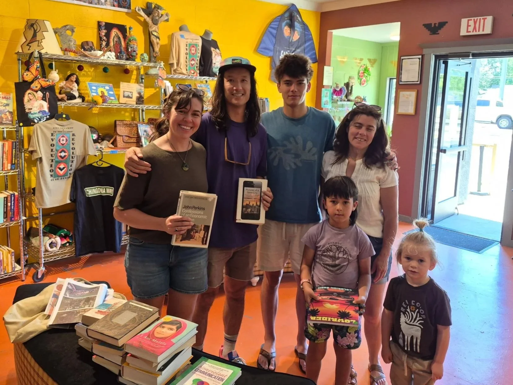 A group of six people, including two children, standing inside a brightly decorated bookshop or library. They are smiling and holding books, with a table of books in the foreground. The background features colorful walls, shelves with various items, and a large window or door letting in natural light.