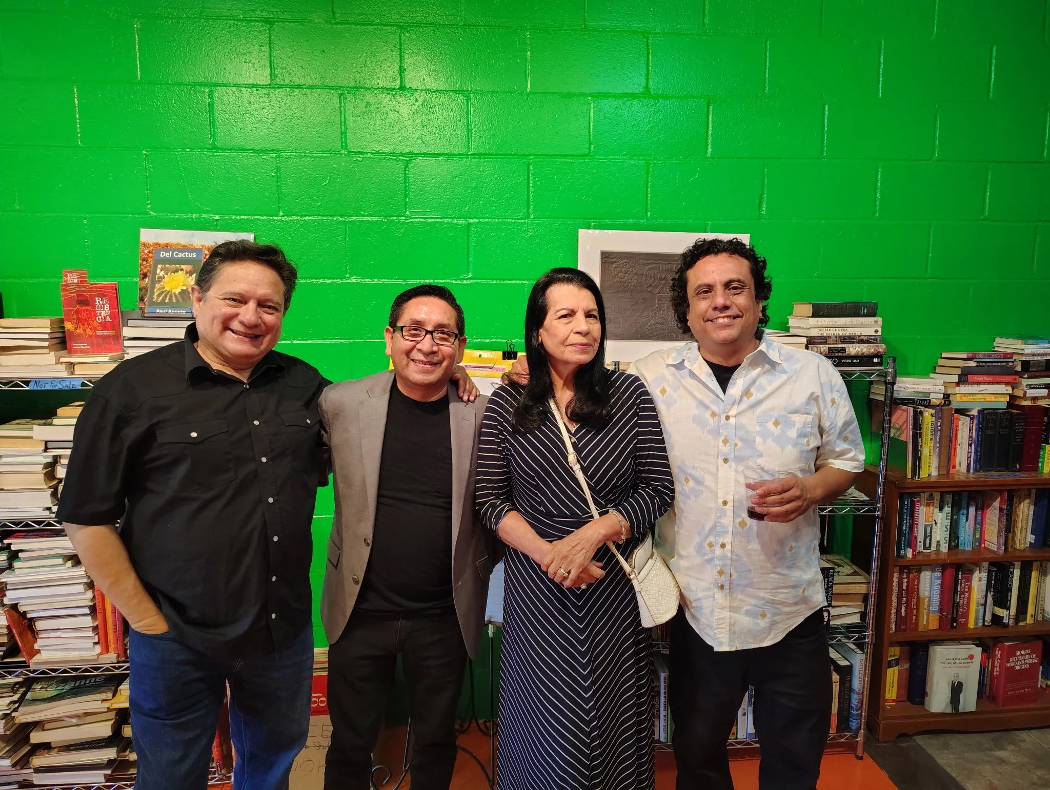 Four individuals standing together in front of green-painted brick wall, with stacks of books on shelves behind them at a bookstore or library event.