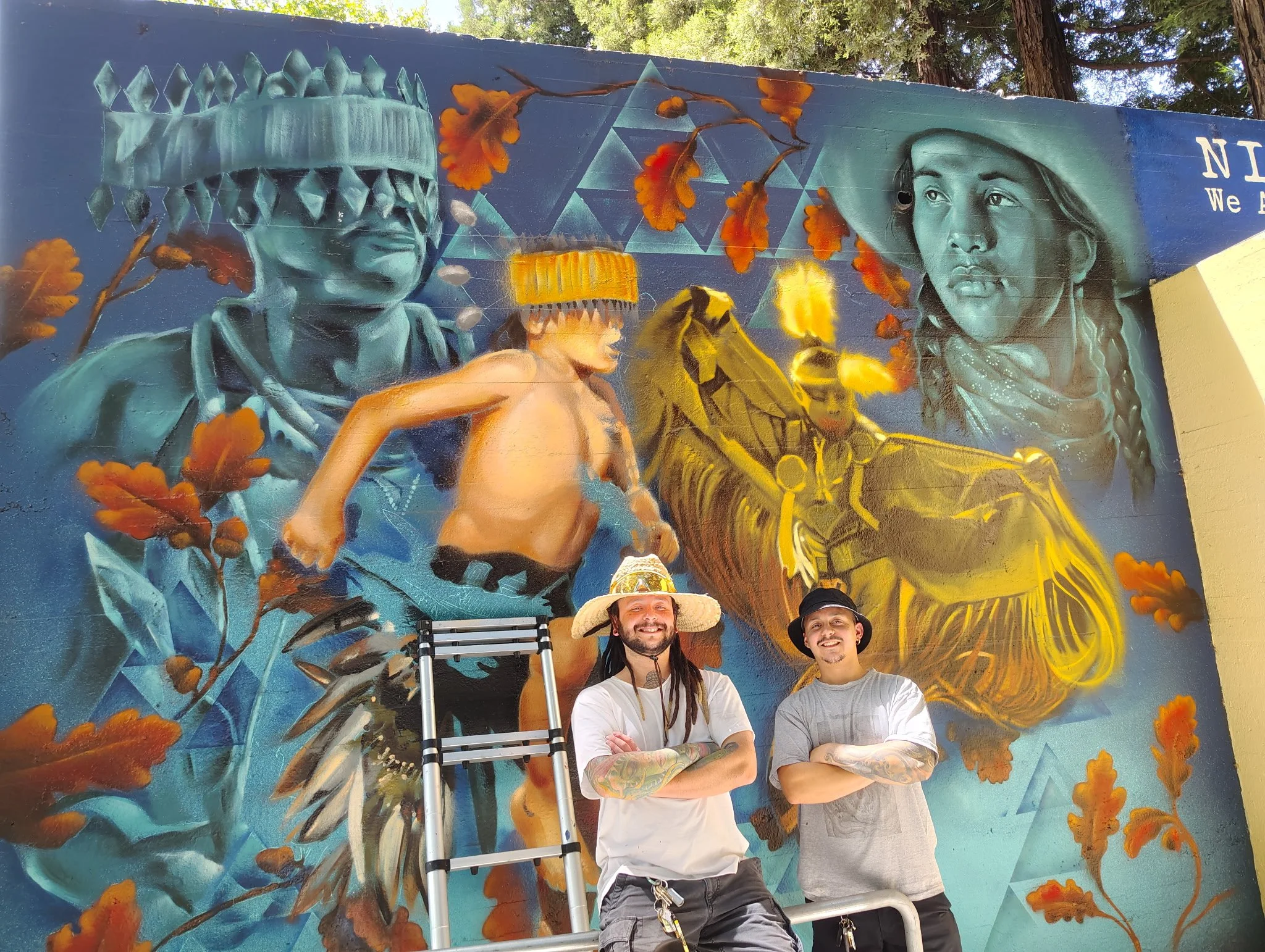 Two men with tattoos and hats standing in front of a vibrant mural painting of indigenous figures, a child, and geometric patterns, outdoors with trees in the background.