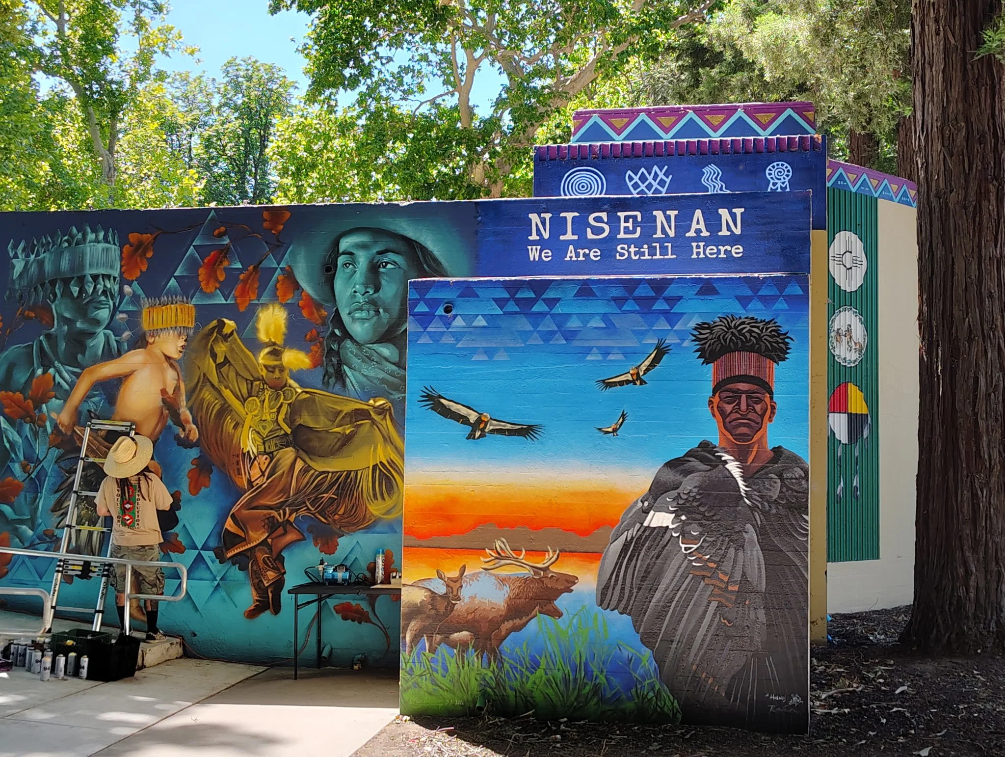 Colorful mural outdoors with the text 'Nisenan We Are Still Here' and portraits of Native American individuals surrounded by nature imagery, trees, and birds.