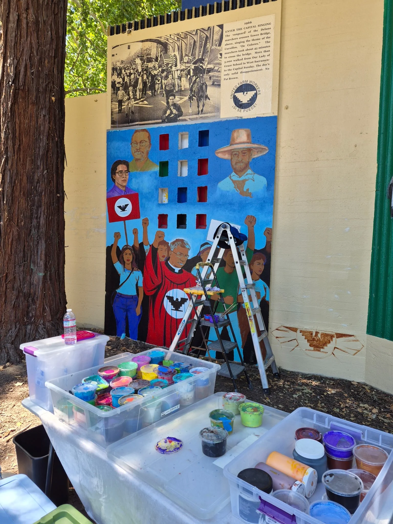 A mural depicting diverse people with raised fists, holding signs and flags, celebrating unity and protest. The scene is colorful and vibrant, emphasizing activism and strength.