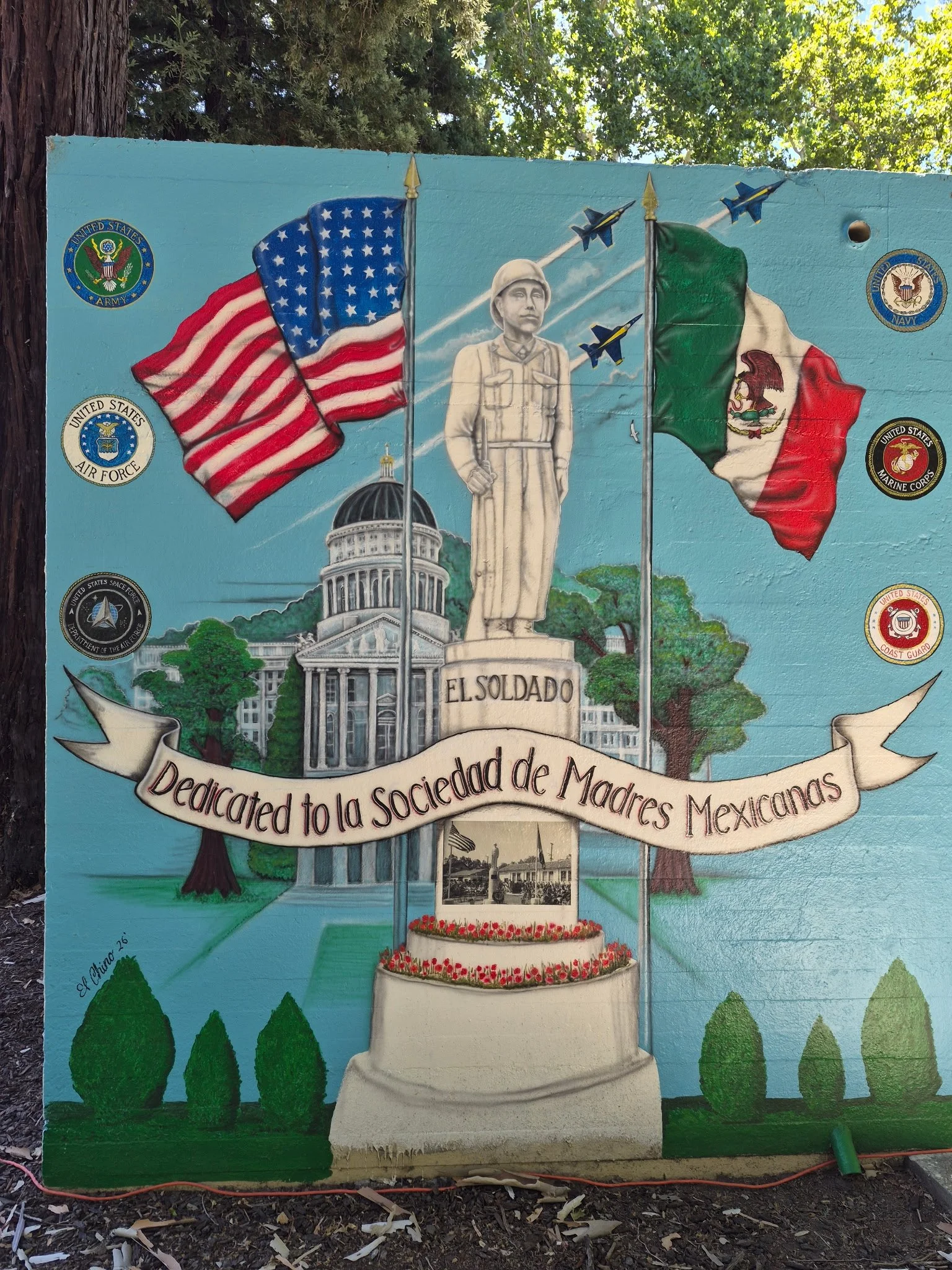 A mural dedicated to Mexican women veterans, showing a statue of a female soldier with flags of the United States and Mexico behind her. The mural includes the seals of various U.S. military branches and a banner with a Spanish inscription. The scene