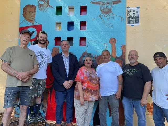 Seven people standing in front of a painted mural on a wall, with some artwork and a small stool visible.