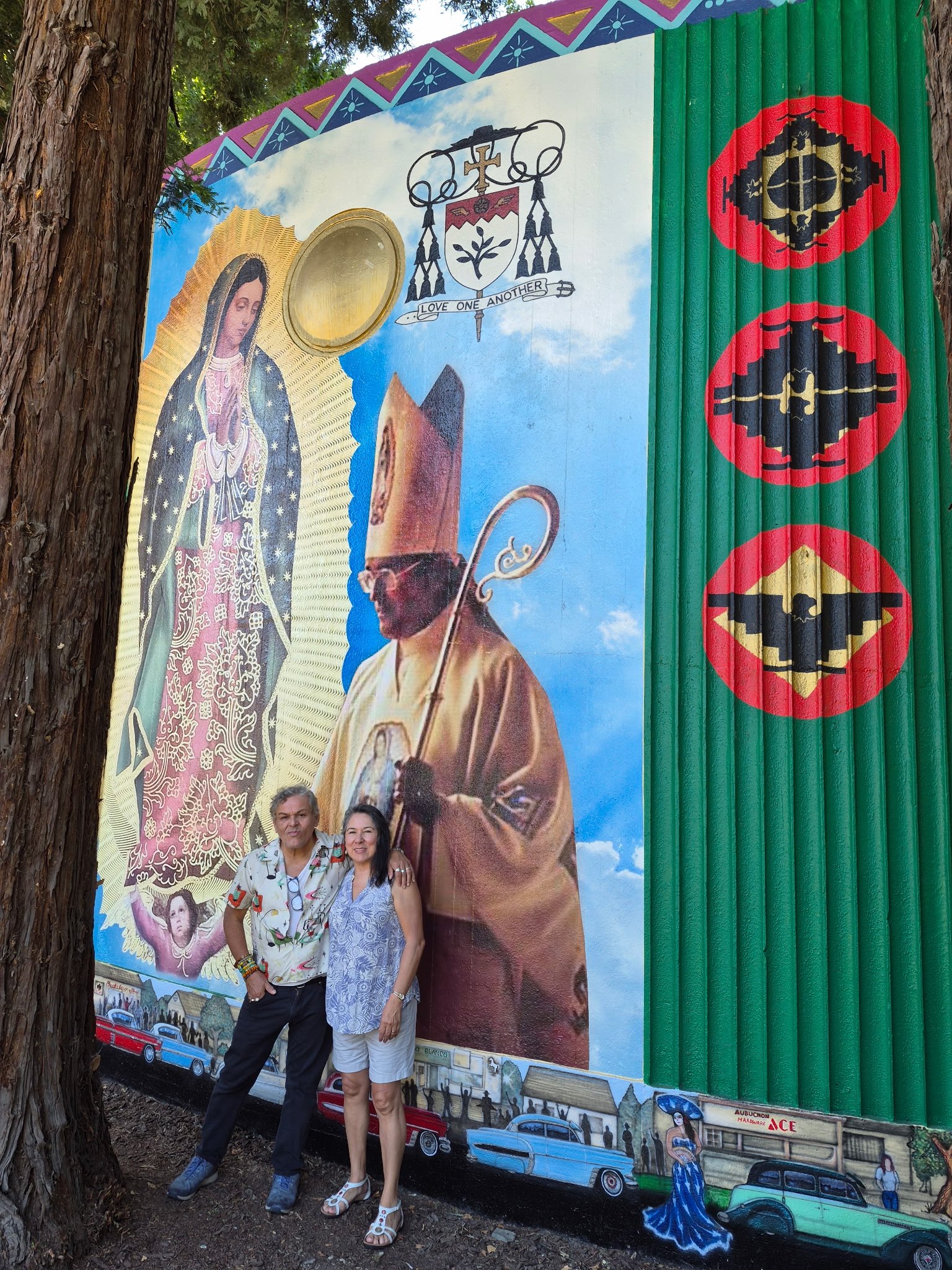Two people, a man and a woman, standing in front of a colorful mural painted on a wall with trees nearby. The mural depicts religious and cultural imagery, including the Virgin Mary, a man in traditional Mexican attire, and various symbols and scenes