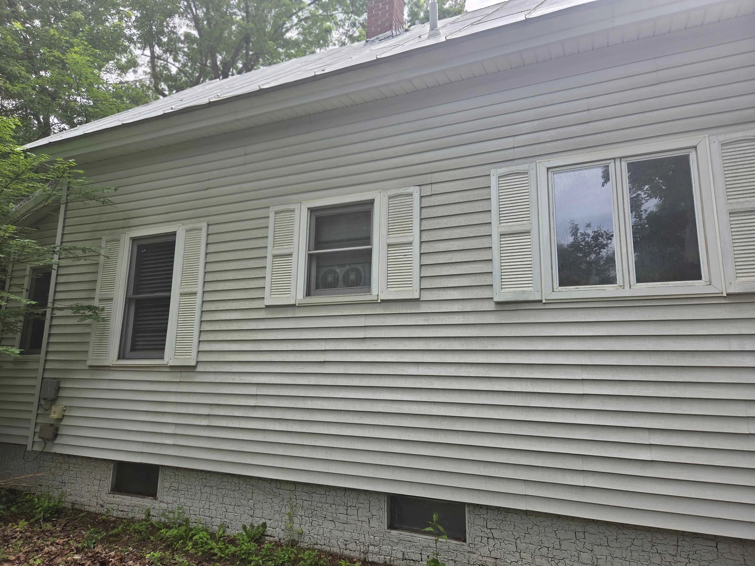 The back of a house with beige vinyl siding, three windows with white shutters, and a concrete foundation with visible cracks. There are some green plants and trees surrounding the house.