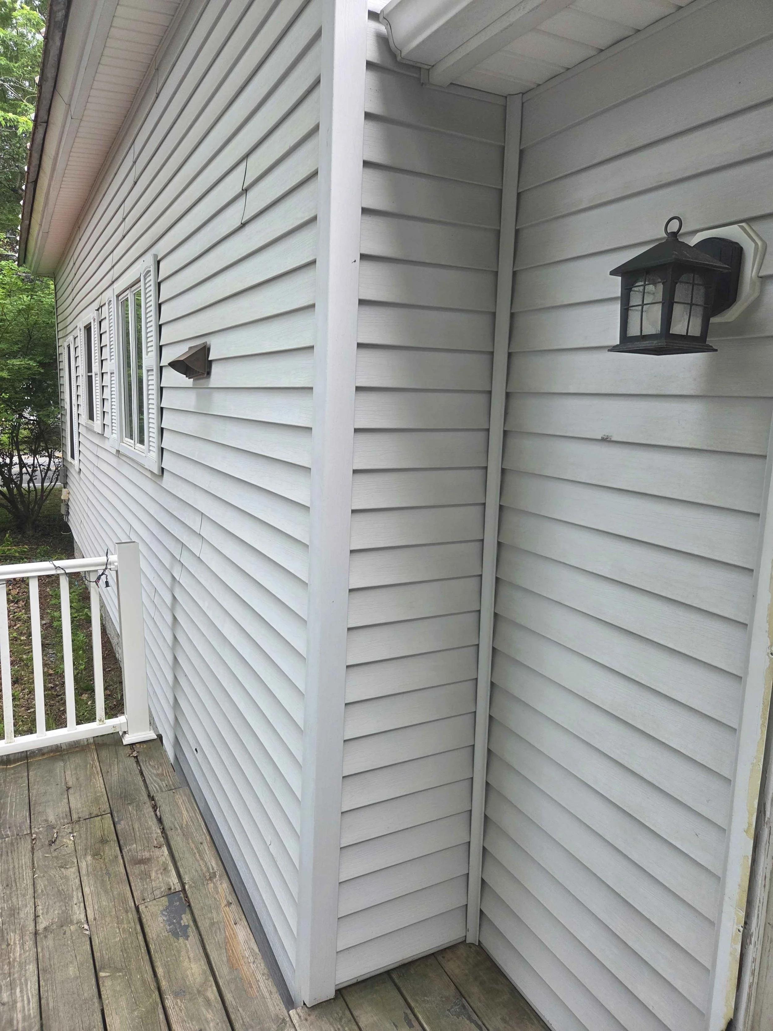View of the side of a house with white vinyl siding, three small windows with shutters, a wall-mounted outdoor lantern, a small wall-mounted light, a white railing, and a wooden deck.
