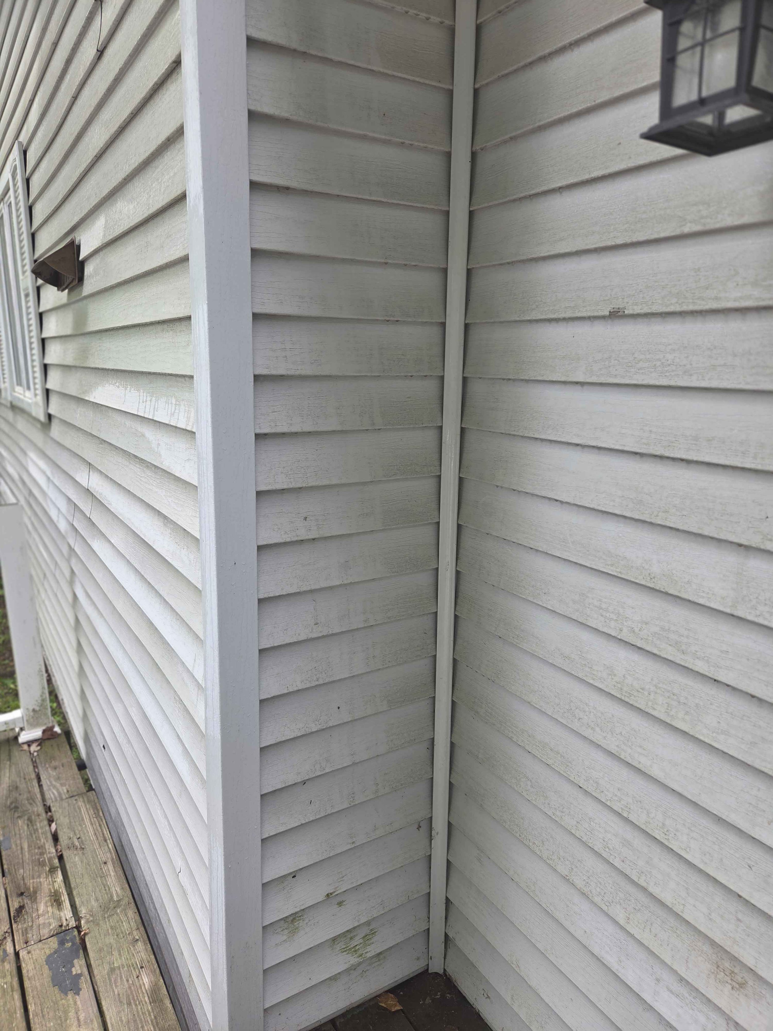 The corner of a house showing white horizontal siding with some weathering and dirt, a gutter on the left side, and a black lantern-style light fixture hanging on the right side.