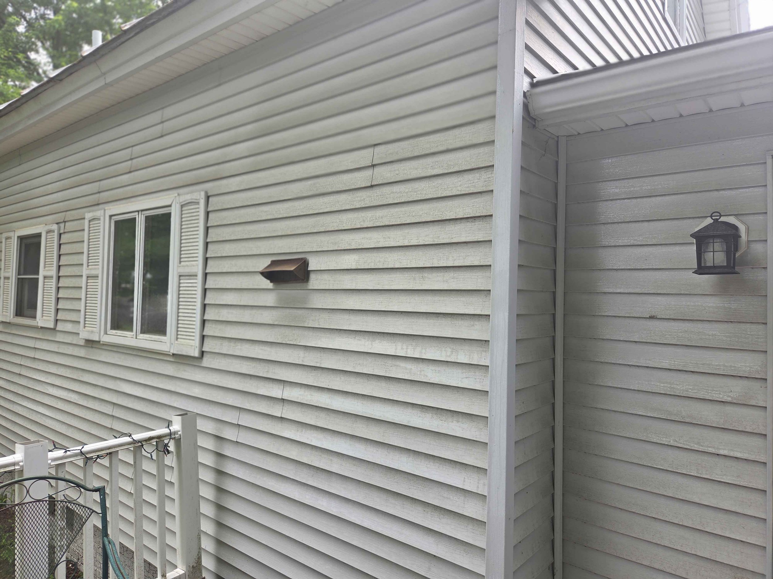 Back of a house with beige vinyl siding, three windows with white shutters, a small brown vent, and a black wall-mounted lantern light. A white railing with string lights is visible in the lower left corner.