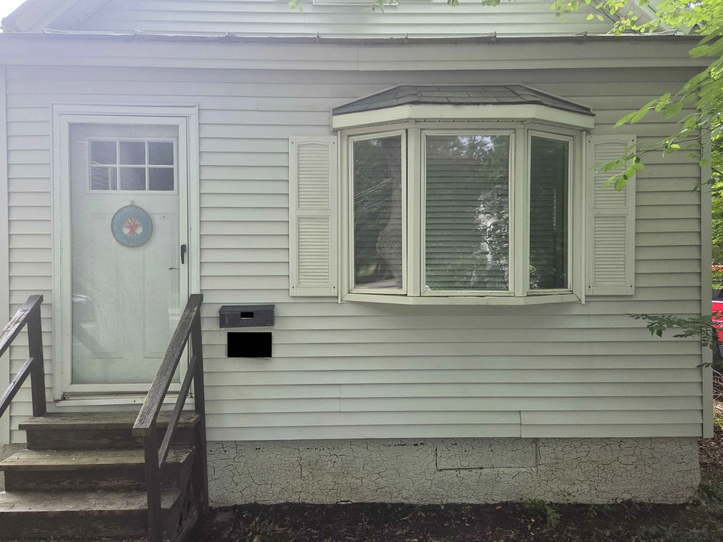 Back of a house with beige siding, a small porch with dark wood stairs, a white door with a decorative wreath, and a large bay window with white shutters.