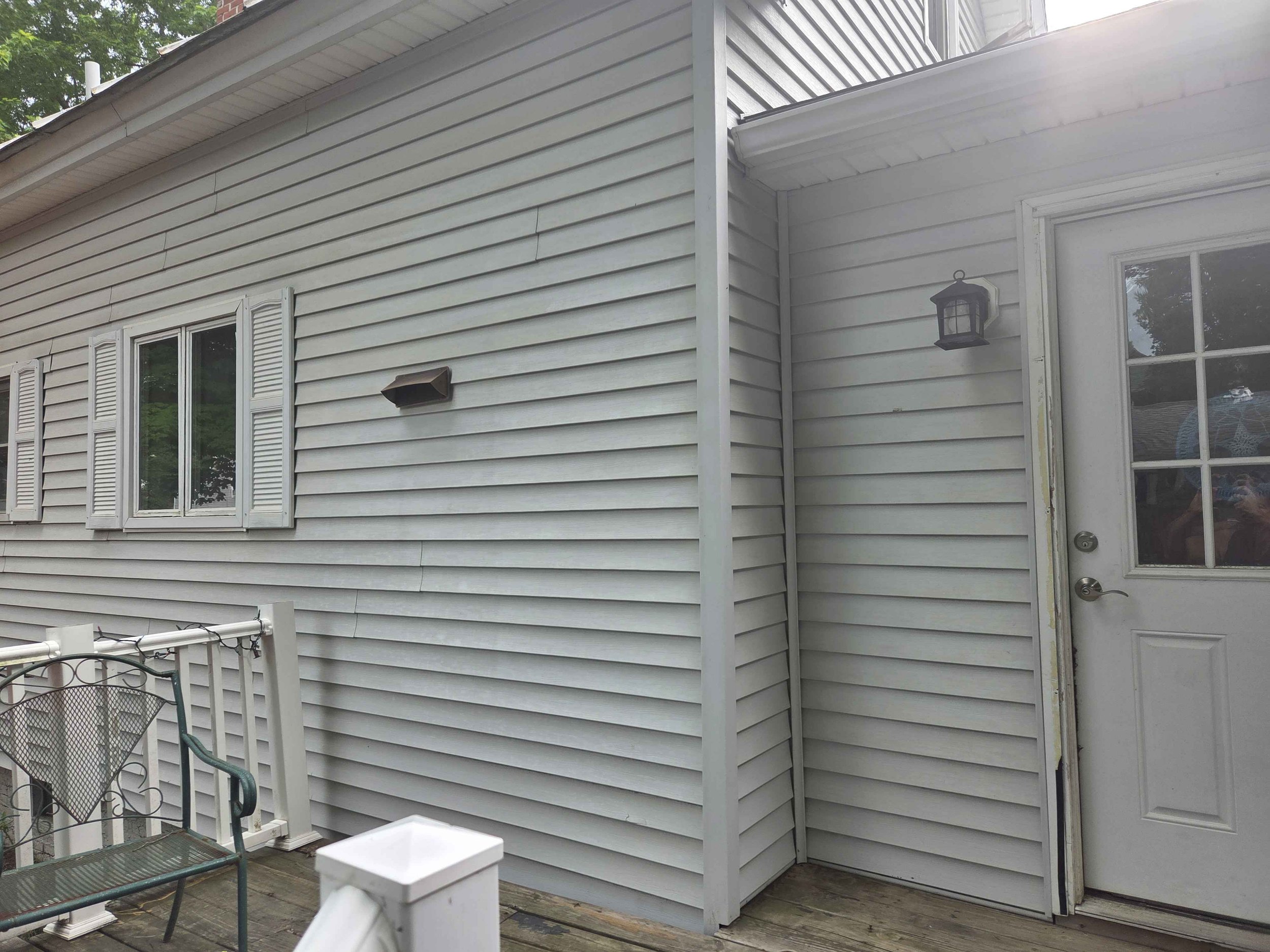White vinyl-sided house exterior with three windows, a door with glass panel, outdoor wall lantern, black outdoor vent, and patio furniture including a metal chair and a small table.