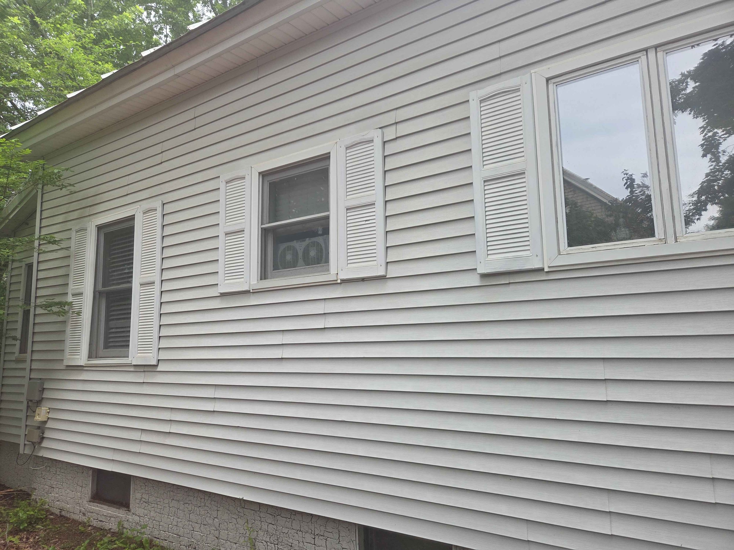 Exterior side view of a house with white vinyl siding, three windows with white shutters, and a basement window near the ground, with some greenery and trees in the background.