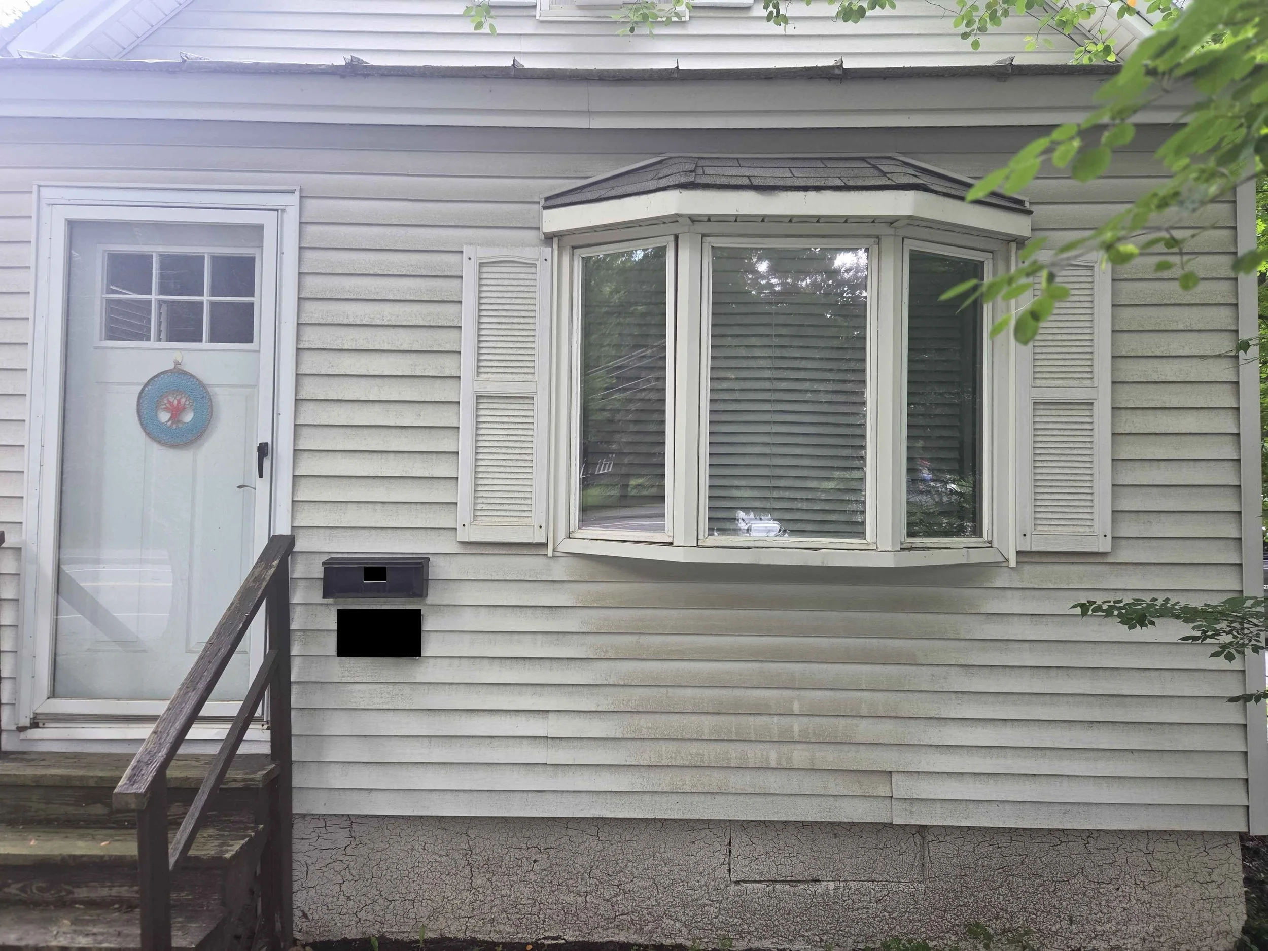 The back exterior of a house with cream-colored vinyl siding, a white door with a red, blue, and teal wreath, a bay window with shutters, a staircase with dark wood railing, and a cracked concrete foundation.