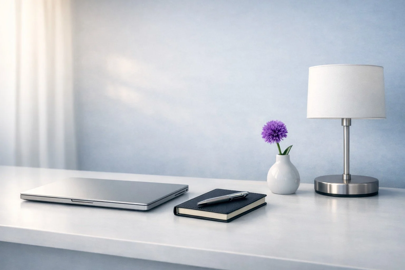 A white desk for Paid Search work with a closed silver laptop, a black notebook with a silver pen on top, a white vase with a purple flower, and a silver and white table lamp against a light blue wall.