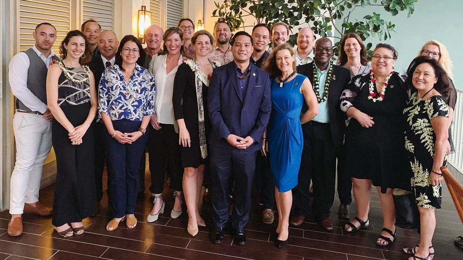 A group of about twenty diverse people dressed in formal and semi-formal attire, posing together indoors in front of a green leafy plant and wall-mounted lights, smiling for a group photo.