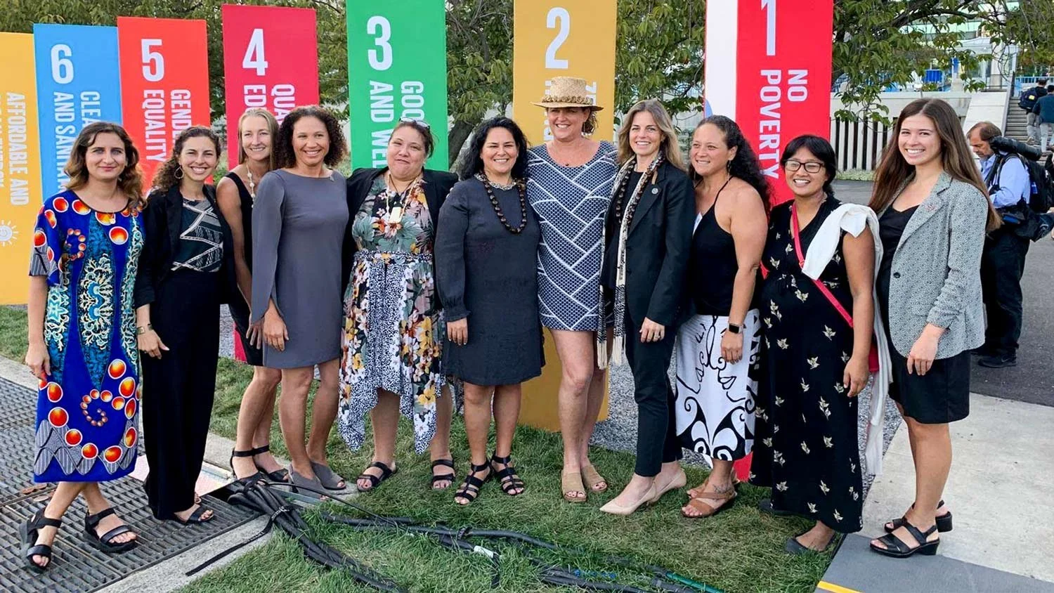 A group of twelve women standing outdoors, smiling, with colorful banners in the background displaying social and political messages including 'No Poverty,' 'Zero Hunger,' 'Good Health and Well-being,' 'Quality Education,' 'Gender Equality,' 'Clean Water and Sanitation,' and 'Affordable and Clean Energy.'