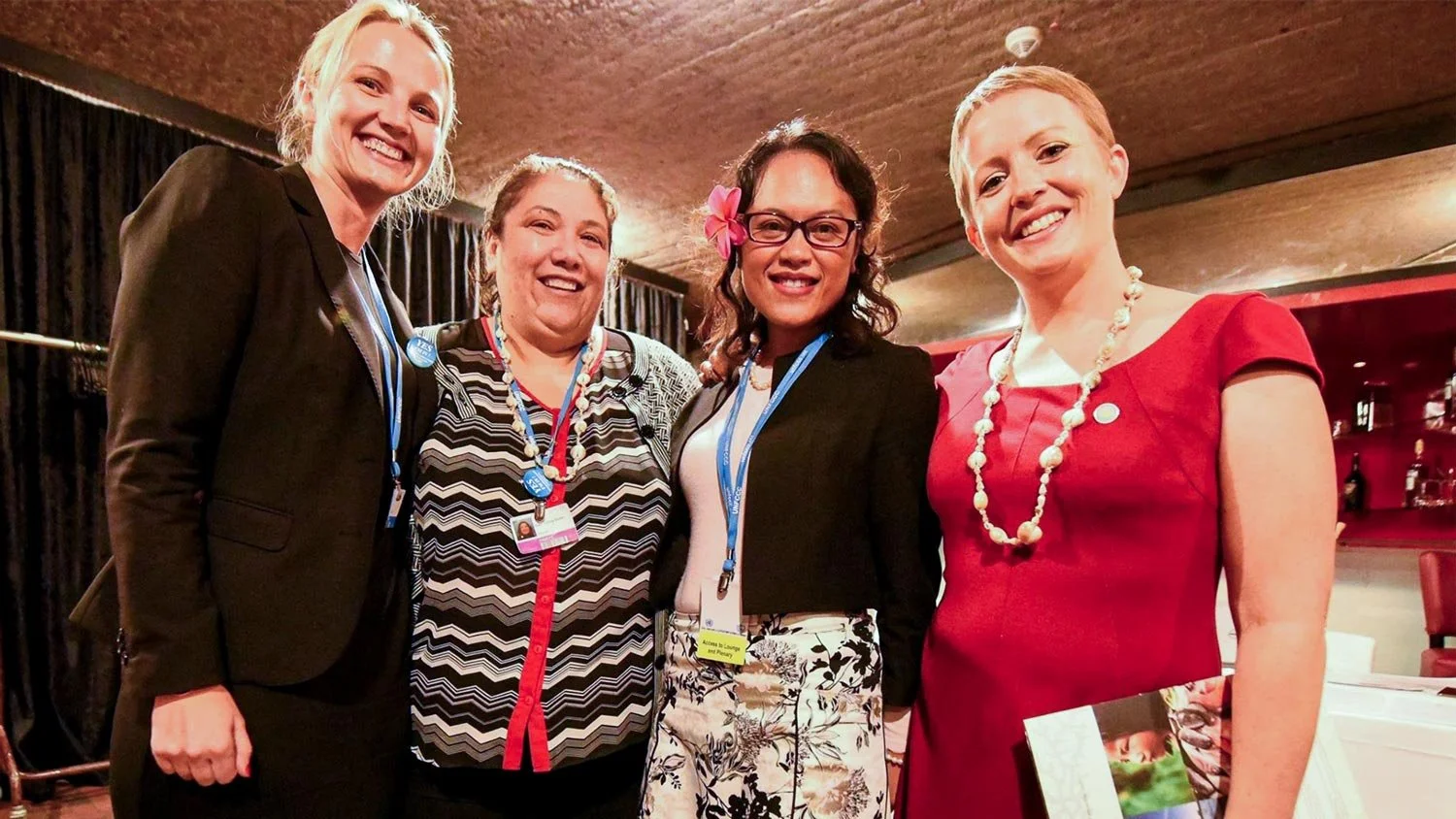 Four women standing close together, smiling for a photo in an indoor setting with a ceiling and wall in the background. They are dressed in business or semi-formal attire.