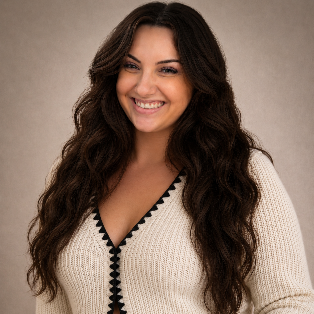 A smiling woman with long, wavy brown hair wearing a cream-colored sweater with black triangular trim, standing against a neutral background.