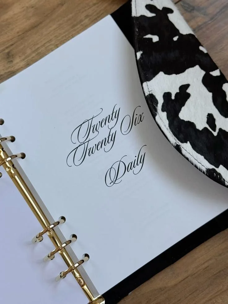 A planner opened to a page with gold rings and cursive writing that says 'Twenty Twenty Six Daily'.