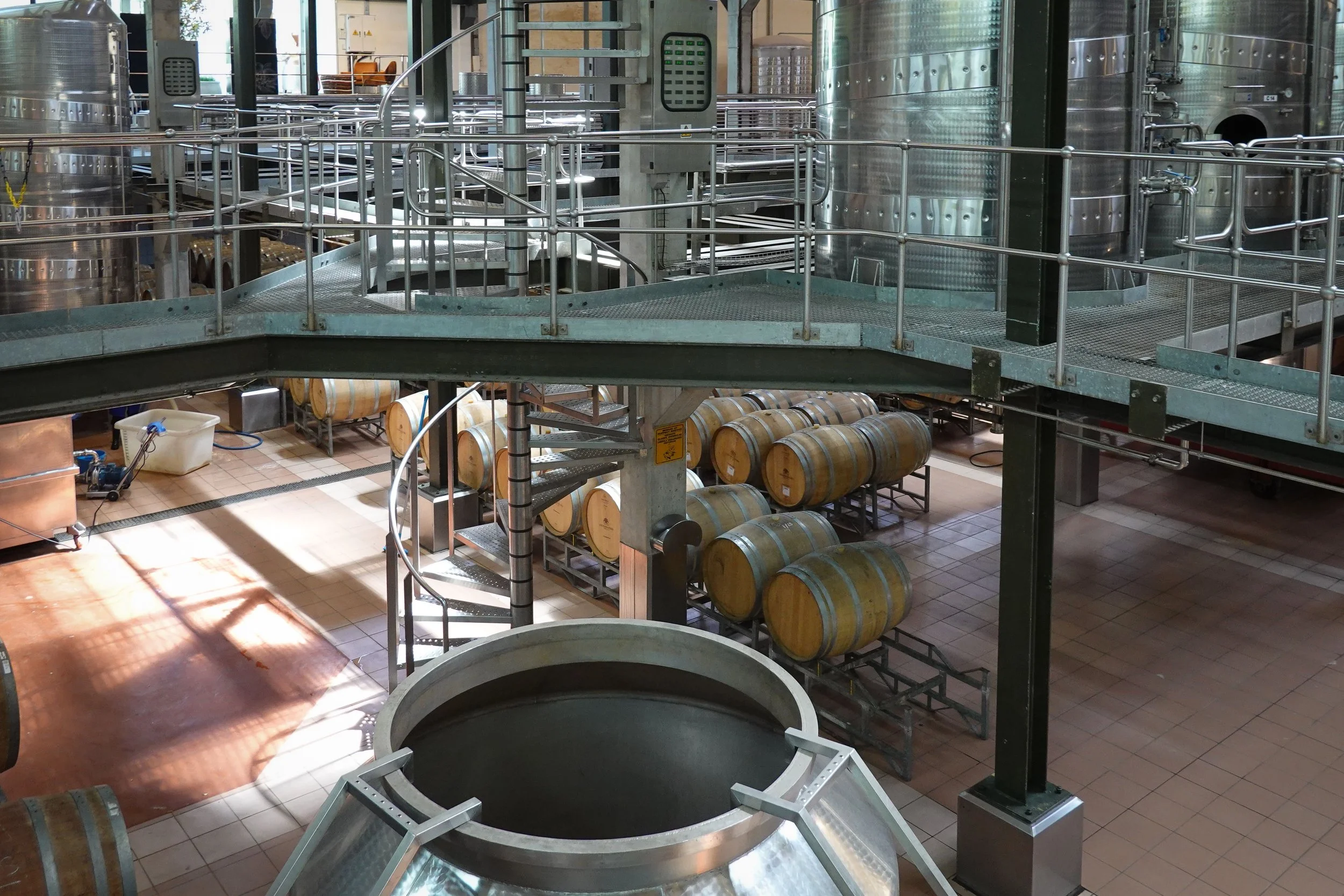 Inside a winery, stainless steel fermentation tanks with metal railings and stairs, wooden barrels stacked underneath, and a tiled floor.